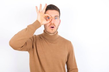 young caucasian man wearing turtleneck over white background doing ok gesture shocked with surprised face, eye looking through fingers. Unbelieving expression.