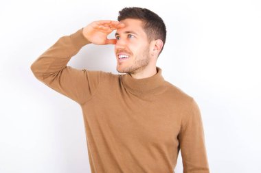 young caucasian man wearing turtleneck over white background very happy and smiling looking far away with hand over head. Searching concept.