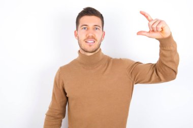 young caucasian man wearing turtleneck over white background smiling and confident gesturing with hand doing small size sign with fingers looking and the camera. Measure concept