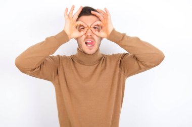 young caucasian man wearing turtleneck over white background doing ok gesture like binoculars sticking tongue out, eyes looking through fingers. Crazy expression.