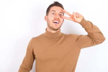 young caucasian man wearing turtleneck over white background Doing peace symbol with fingers over face, smiling cheerful showing victory
