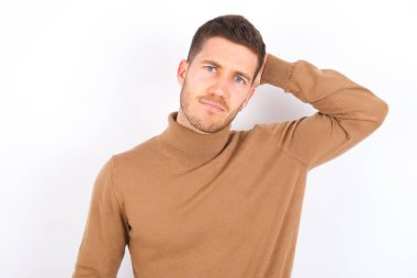 young caucasian man wearing turtleneck over white background being confused and wonders about something. Holding hand on head, uncertain with doubt. Pensive concept.