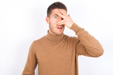 young caucasian man wearing turtleneck over white background peeking in shock covering face and eyes with hand, looking through fingers with embarrassed expression.