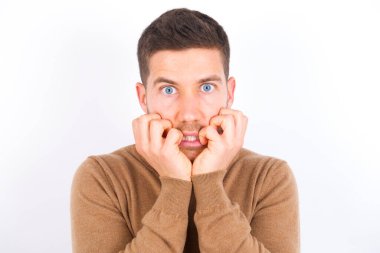 Fearful young caucasian man wearing turtleneck over white background keeps hands near mouth, feels frightened and scared,  has a phobia,  Shock and frighted concept.