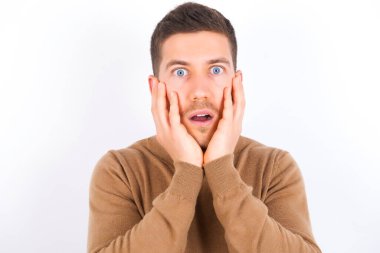 Stupefied young caucasian man wearing turtleneck over white background expresses excitement and thrill, keeps jaw dropped, hands on cheeks, has eyes popped out