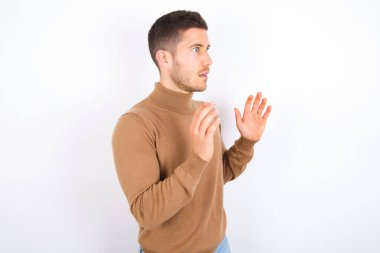 young caucasian man wearing turtleneck over white background shouts loud, keeps eyes opened and hands tense.