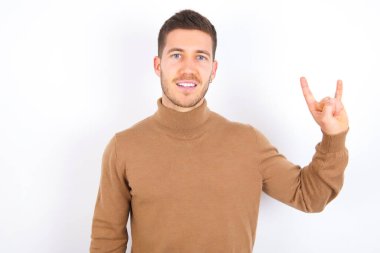 young caucasian man wearing turtleneck over white background doing a rock gesture and smiling to the camera. Ready to go to her favorite band concert.