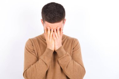 young caucasian man wearing turtleneck over white background covering her face with her hands, being devastated and crying. Sad concept