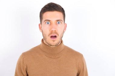 Oh my God. Surprised young caucasian man wearing turtleneck over white background stares at camera with shocked expression exclaims with unexpectedness,