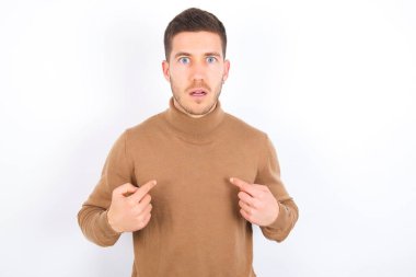Embarrassed young caucasian man wearing turtleneck over white background indicates at herself with puzzled expression, being shocked to be chosen to participate in competition, hesitates about something