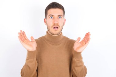 Surprised terrified young caucasian man wearing turtleneck over white background Gestures with uncertainty, stares at camera, puzzled as doesn't know answer on tricky question, People, body language, emotions concept