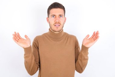 Indignant young caucasian man wearing turtleneck over white background gestures in bewilderment, frowns face with dissatisfaction.