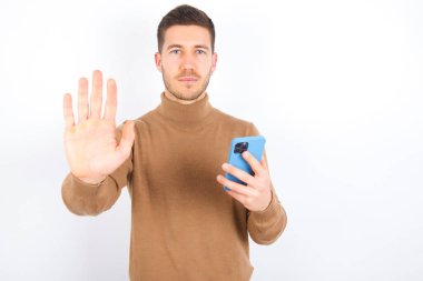 young caucasian man wearing turtleneck over white background using and texting with smartphone with open hand doing stop sign with serious and confident expression, defense gesture
