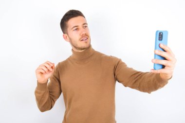 young caucasian man wearing turtleneck over white background smiling and taking a selfie ready to post it on her social media.
