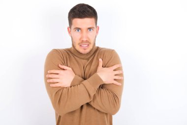 Desperate young caucasian man wearing turtleneck over white background trembles and feels cold, hugs oneself to warm up or feels scared notices something terrifying.
