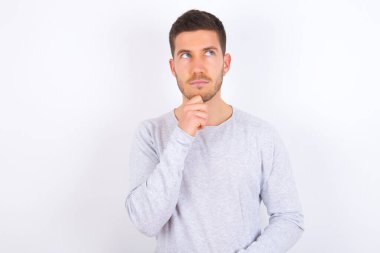 Beyaz arka plan üzerine günlük kıyafetler giyen düşünceli genç beyaz bir adam çenesini tutup kafasını çeviriyor ve harika bir plan yapıyor.