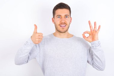 young caucasian man wearing casual clothes over white background smiling and looking happy, carefree and positive, gesturing victory or peace with one hand