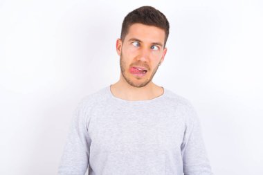 Funny young caucasian man wearing casual clothes over white background makes grimace and crosses eyes plays fool has fun alone sticks out tongue.