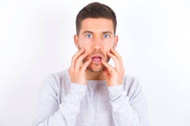 Speechless young caucasian man wearing casual clothes over white background keeps hands near opened mouth reacts to shocking news stares wondered at camera