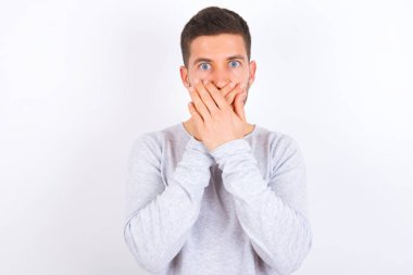 Stunned young caucasian man wearing casual clothes over white background covers both hands on mouth, afraids of something astonishing
