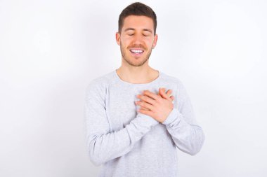 young caucasian man wearing casual clothes over white background expresses happines, laughs pleasantly, keeps hands on heart