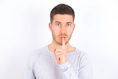 Surprised young caucasian man wearing casual clothes over white background makes silence gesture, keeps finger over lips and looks mysterious at camera