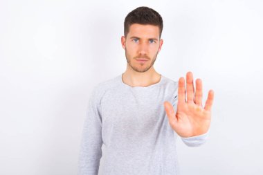 young caucasian man wearing casual clothes over white background shows stop sign prohibition symbol keeps palm forward to camera with strict expression