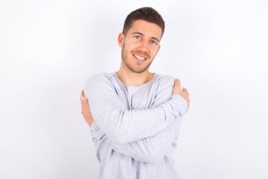 Charming pleased young caucasian man wearing casual clothes over white background embraces own body, pleasantly feels comfortable poses. Tenderness and self esteem concept