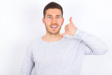 young caucasian man wearing casual clothes over white background makes phone gesture, says call me back again, has glad expression.