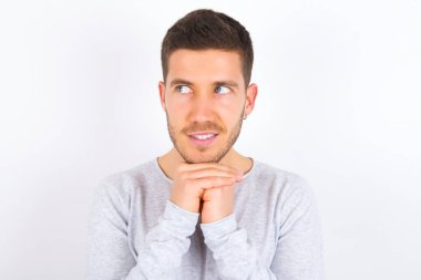 Happy young caucasian man wearing casual clothes over white background anticipates something awesome happen, looks happily aside, keeps hands together near face, has glad expression.