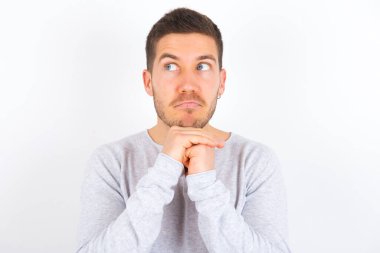 Curious young caucasian man wearing casual clothes over white background keeps hands under chin bites lips and looks with interest aside.
