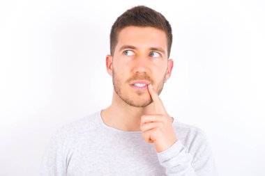 Lovely dreamy young caucasian man wearing casual clothes over white background keeps finger near lips looks aside copy space.