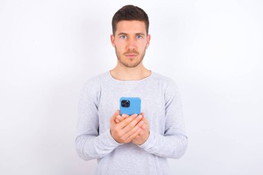 Portrait of serious confident young caucasian man wearing casual clothes over white background holding phone in two hands