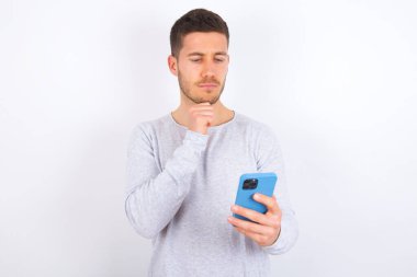 Thoughtful happy young caucasian man wearing casual clothes over white background hold look phone, copyspace