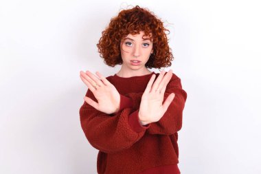 young caucasian woman red haired wearing red sweater over white background keeps has rejection expression crossing arms and palms doing negative sign, angry face.