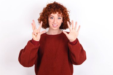 young caucasian woman red haired wearing red sweater over white background keeps showing and pointing up with fingers number nine while smiling confident and happy.