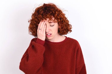 young caucasian woman red haired wearing red sweater over white background keeps Yawning tired covering half face, eye and mouth with hand. Face hurts in pain.