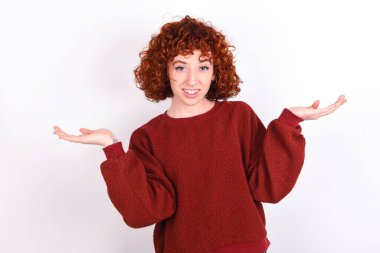 Cheerful cheery optimistic young caucasian woman red haired wearing red sweater over white background keeps holding two palms copy space