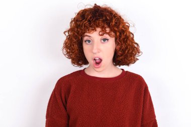 Oh my God. Surprised young caucasian woman red haired wearing red sweater over white background keeps stares at camera with shocked expression exclaims with unexpectedness,