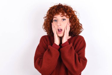 Scared terrified young caucasian woman red haired wearing red sweater over white background keeps shocked with prices at shop, People and human emotions concept