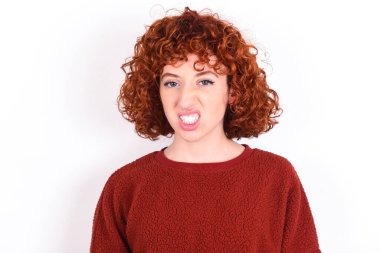young caucasian woman red haired wearing red sweater over white background keeps keeps teeth clenched, frowns face in dissatisfaction, irritated because of much duties.