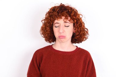 Dismal gloomy rejected young caucasian woman red haired wearing red sweater over white background keeps has problems and difficulties, curves lower lip and closes eyes in despair, being in depression