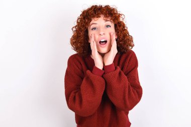 young caucasian woman red haired wearing red sweater over white background keeps shouting excited to front.