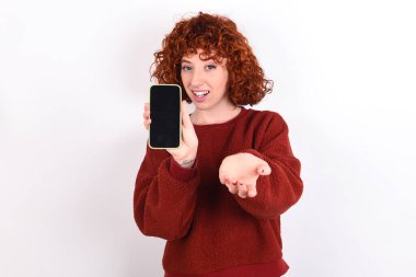 young caucasian woman red haired wearing red sweater over white background keeps with a mobile. presenting smartphone. Advertisement concept.