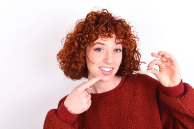 Beyaz arka plan üzerinde kırmızı kazak giyen beyaz saçlı genç beyaz kadın görünmez bir hizalama tutuyor ve mükemmel düz dişlerini işaret ediyor. Diş sağlığı ve güven kavramı.