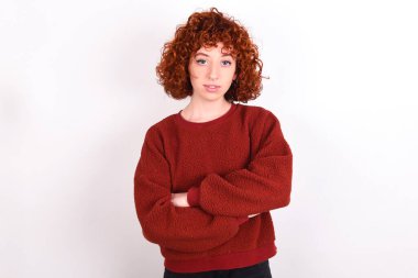 Self confident serious calm young caucasian woman red haired wearing red sweater over white background keeps stands with arms folded. Shows professional vibe stands in assertive pose.