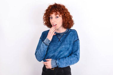 young caucasian woman red haired wearing blue T-shirt over white background covers mouth and looks with wonder at camera, cannot believe unexpected rumors.