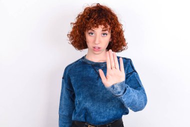 young caucasian woman red haired wearing blue T-shirt over white background shows stop sign prohibition symbol keeps palm forward to camera with strict expression