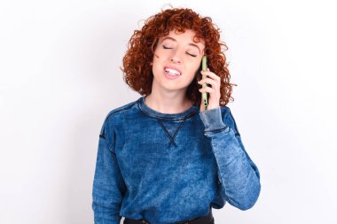Funny young caucasian woman red haired wearing blue T-shirt over white background laughs happily, has phone conversation, being amused by friend, closes eyes.