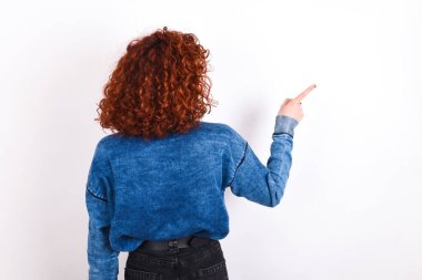 young caucasian woman red haired wearing blue T-shirt over white background pointing to object on copy space, rear view. Turn your back